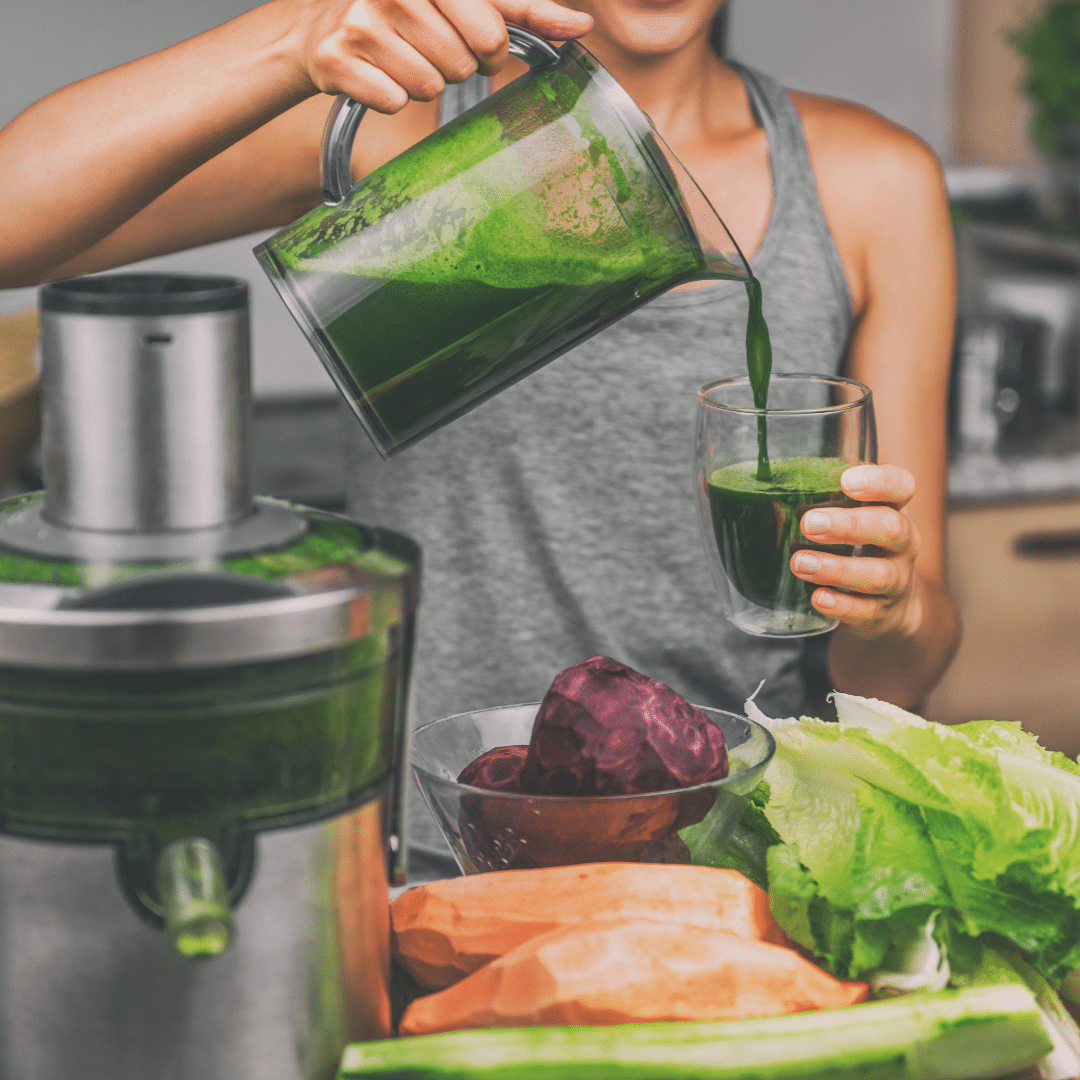 Woman pouring green juice into a glass next to a juicer and fresh vegetables like beetroot.